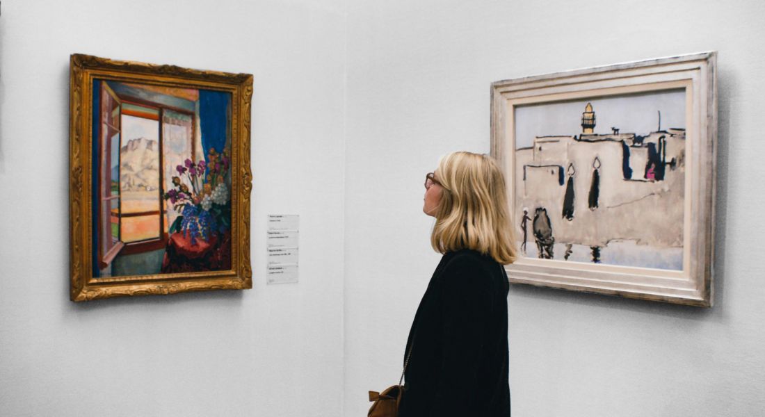 225 Sycamore person with shoulder-length blonde hair, dressed in a black coat, stands in an art gallery viewing two framed paintings on a white wall. One painting depicts a vibrant scene with flowers and an open window, while the other features a more muted, possibly architectural scene.