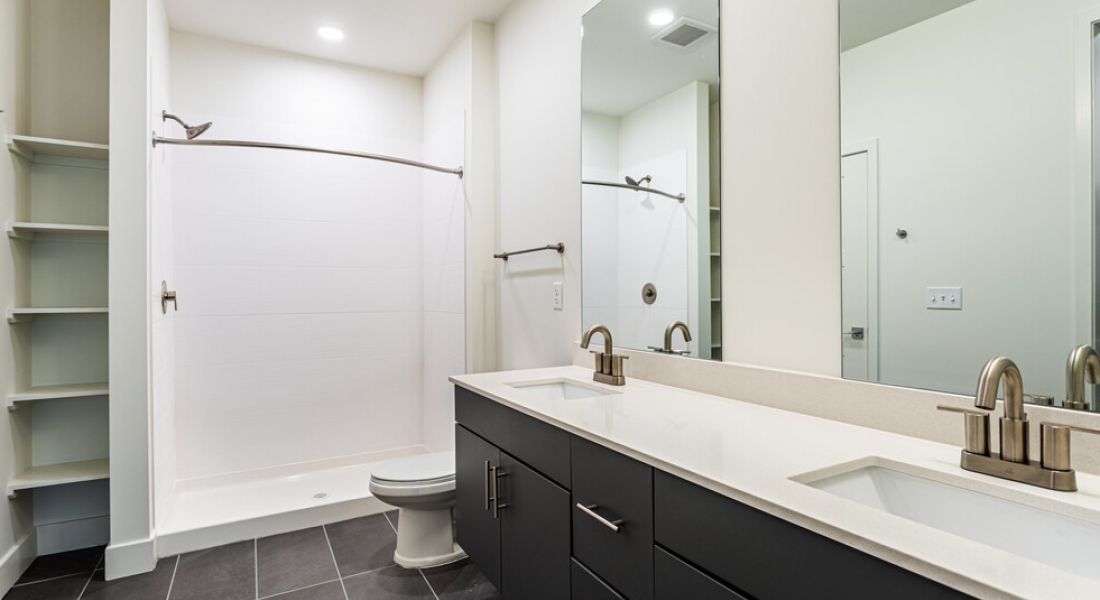 225 Sycamore modern bathroom featuring a large mirror above a double sink vanity with black cabinets and a beige countertop. To the left, there is a spacious, open shower area with a curved shower rod and a white toilet nearby. The floor is tiled in dark gray.