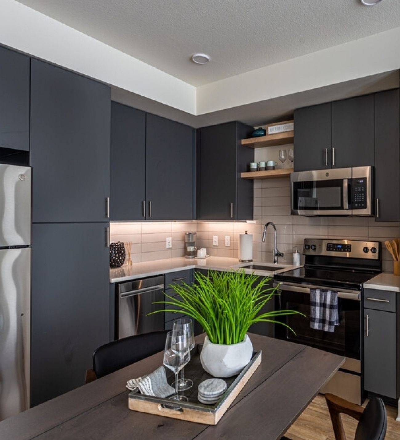 225 Sycamore Modern kitchen with dark cabinets, stainless steel appliances, and under-cabinet lighting. A small potted plant and two glasses on a tray sit on a kitchen table in the foreground. Shelves with various items are above the countertop. Bright and neatly organized space.