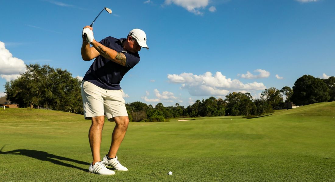 225 Sycamore person wearing a navy shirt, beige shorts, white cap, and white shoes is captured mid-swing on a golf course. The sky is clear with a few clouds, and the green stretches out with trees in the background.