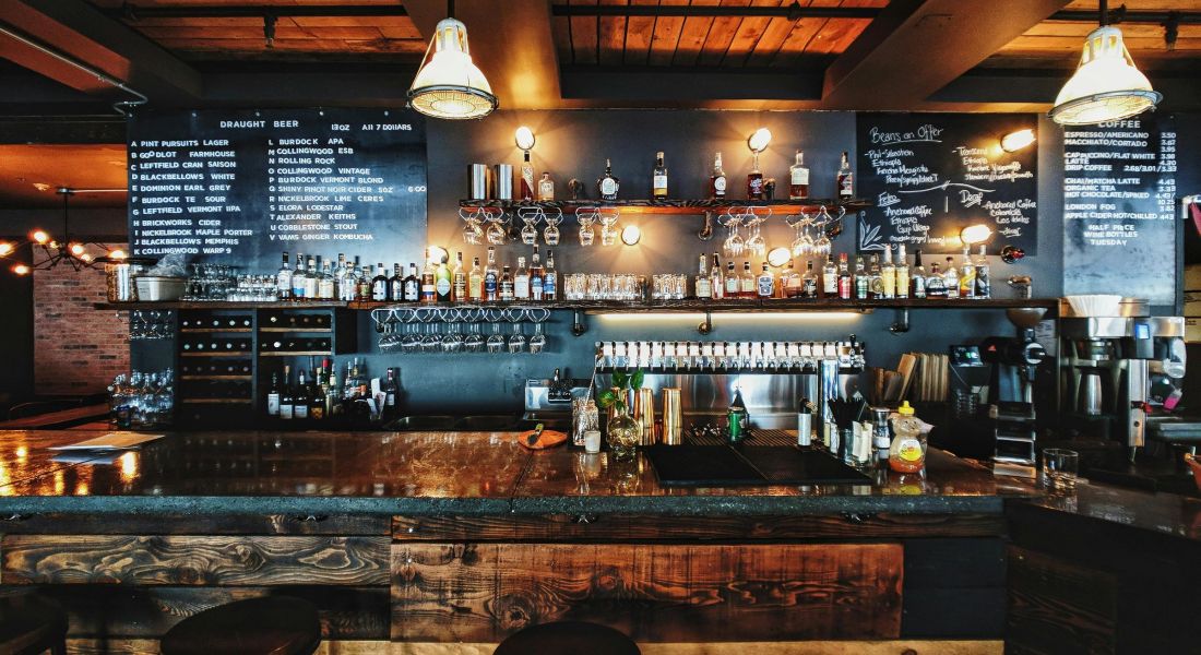 225 Sycamore cozy bar with a rustic wooden counter and two barstools in front. The shelves behind the bar are lined with various bottles of alcohol, and the wall menu displays drink options in white lettering. Warm lighting creates a welcoming atmosphere.