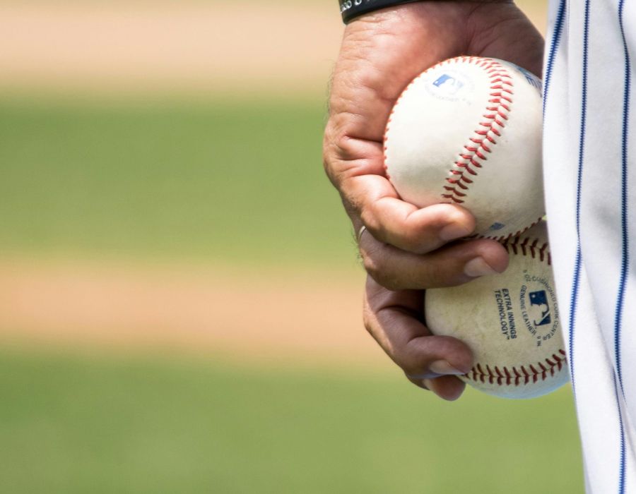 Close-up of a person holding two baseballs in their hand. The background is a blurred baseball field, with green grass and a dirt path visible. The person is wearing a white uniform with a dark wristband on their left wrist.