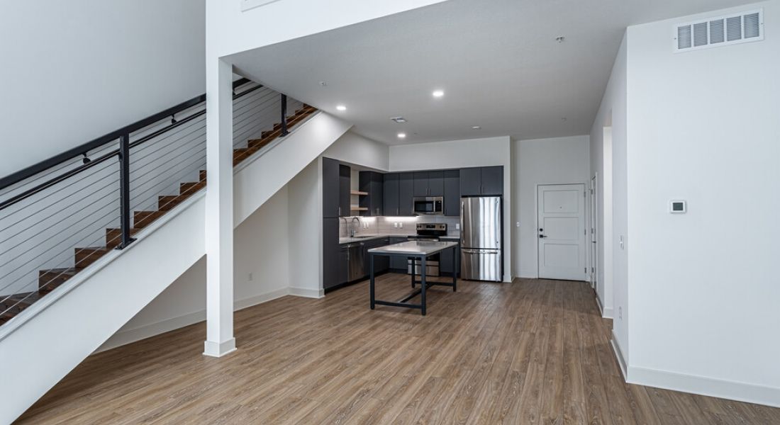 225 Sycamore Modern open-concept kitchen and living area with light wood flooring. The kitchen features dark cabinets, a refrigerator, and an oven, and includes an island with seating. Adjacent is a staircase with a black handrail leading to an upstairs area.