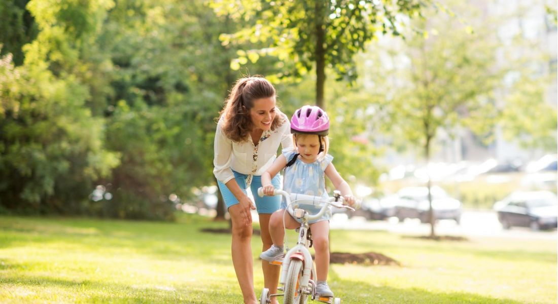 225 Sycamore woman is helping a child learn to ride a bike in a sunny, green park. The child is wearing a pink helmet and smiling, while the woman runs alongside, offering support. Trees and grass surround them, creating a cheerful outdoor setting.