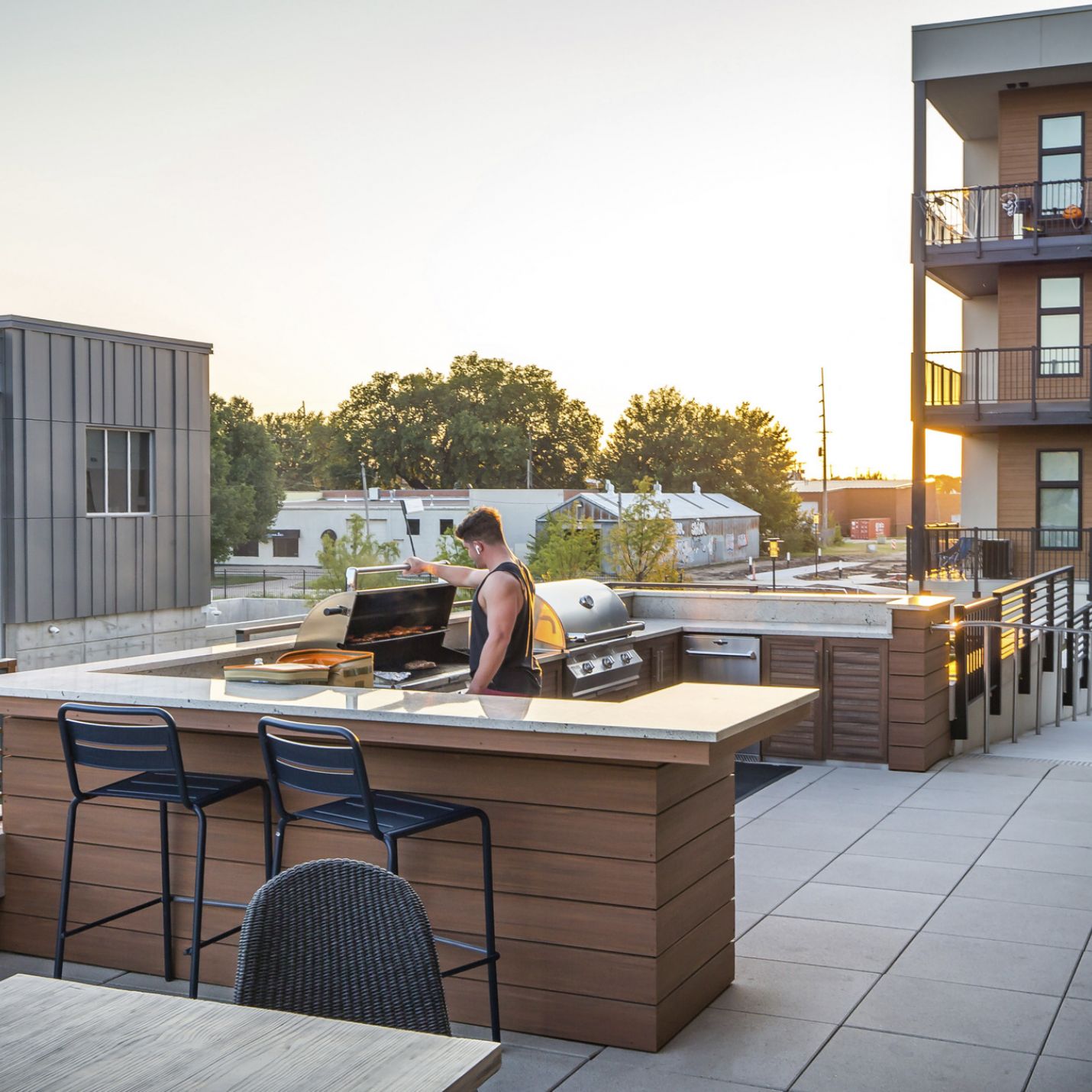 225 Sycamore resident barbequing at a modern outdoor rooftop kitchen area with seating and countertops. The rooftop overlooks a cityscape with trees and buildings visible in the background during sunset.