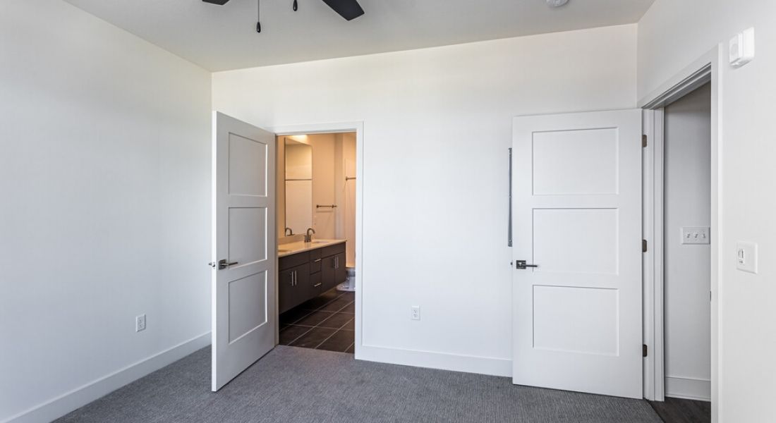225 Sycamore vacant room with white walls and dark grey carpet. The room has two closed white doors on one side and one partially open door revealing a bathroom sink and vanity with mirror. A ceiling fan with light is mounted on the white ceiling.
